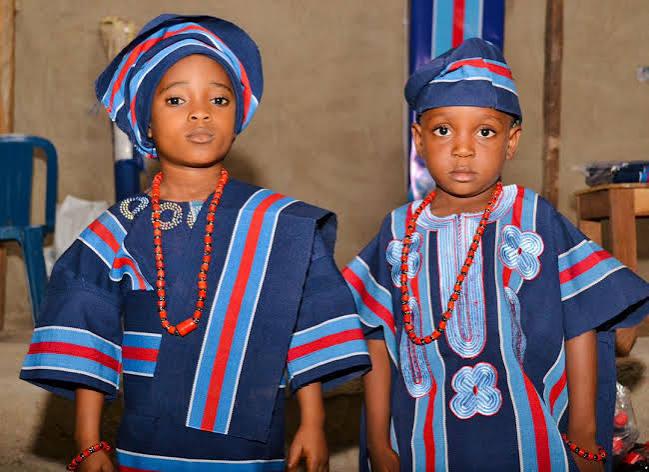 Two children in traditional Takete-Ide attire, wearing beaded necklaces and matching caps