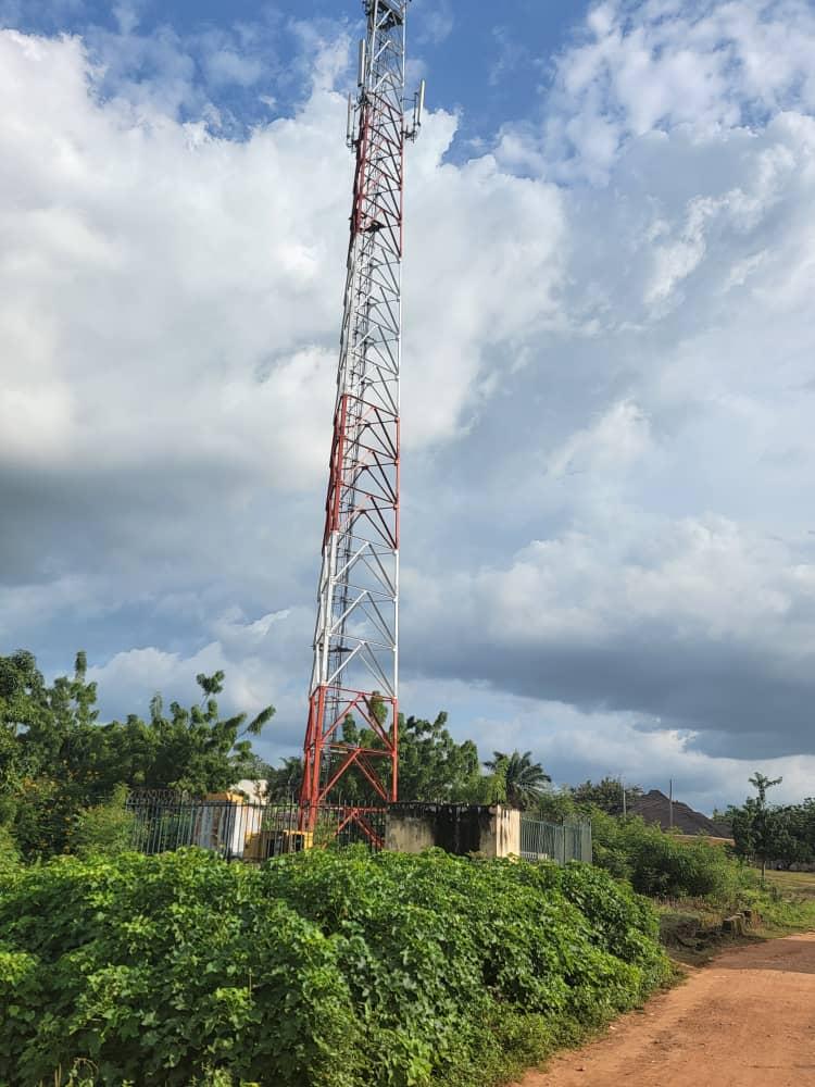 A red and white telecommunications mast standing above green vegetation beside a dirt road